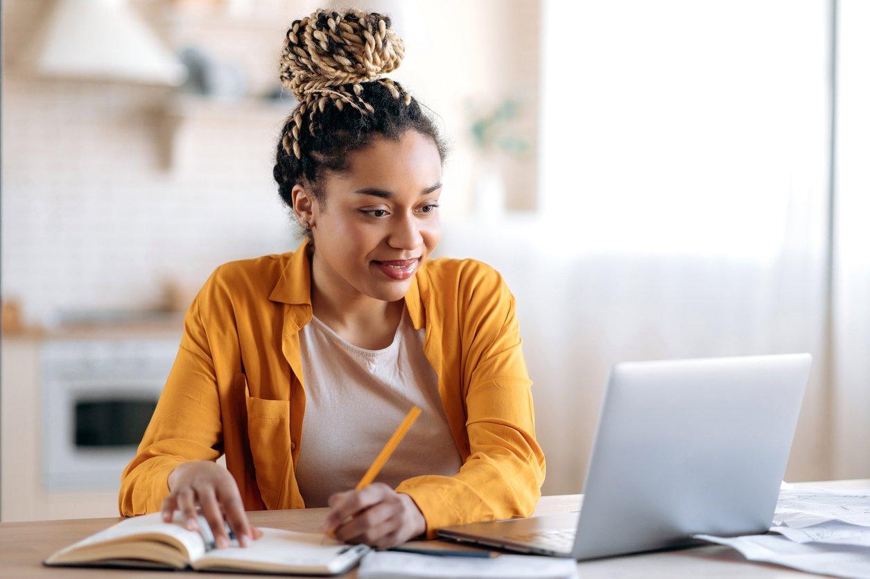 Focado elegante estudante afro-americana com dreadlocks afro, estudando remotamente de casa, usando um laptop, tomando notas no bloco de notas durante a aula online, conceito de e-learning, sorrindo.