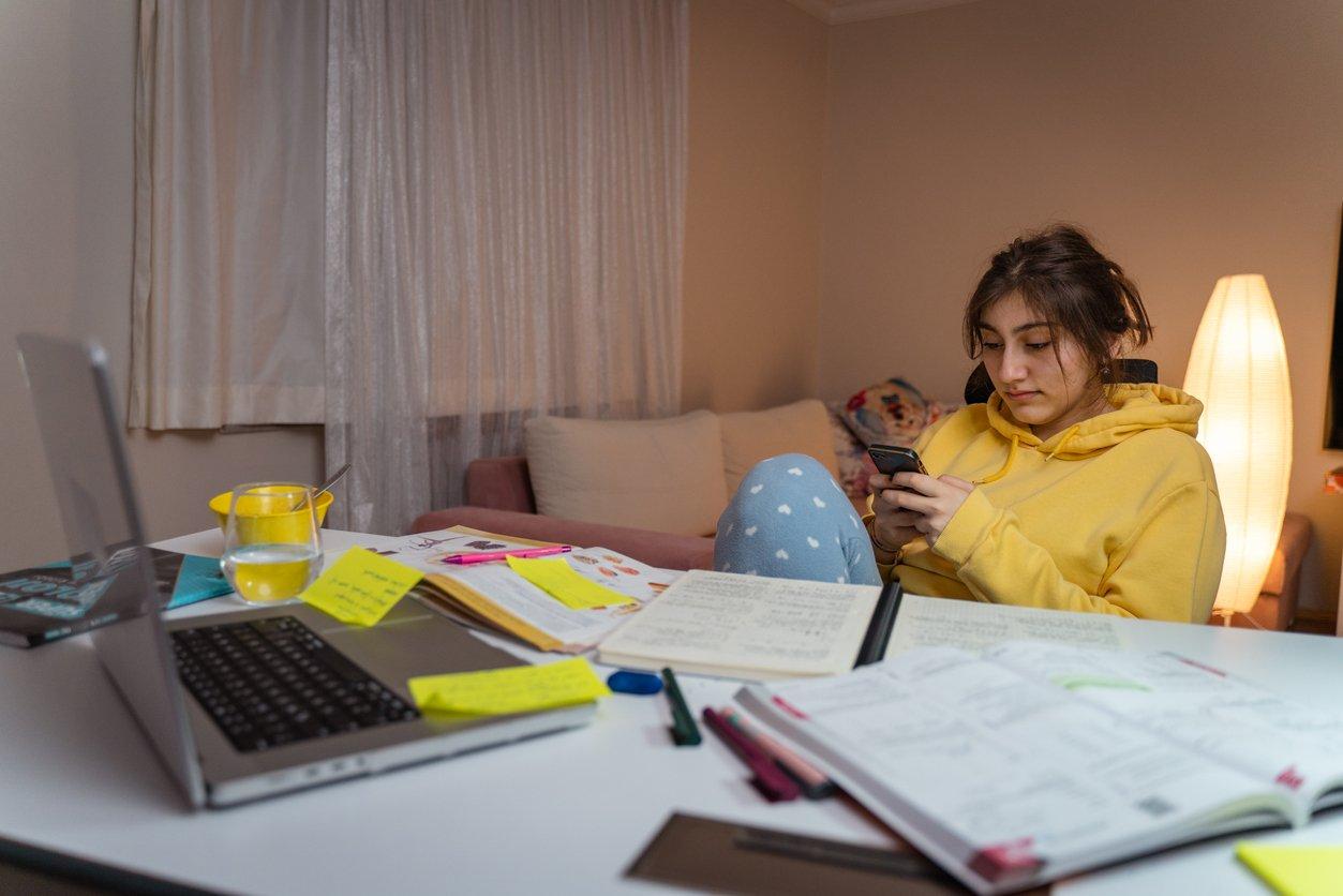 Menina de casaco amarelo sentada em uma cadeira com o celular na mão. à sua frente uma mesa cheia de livros abertos. A imagem representa que ela está exercendo a procrastinação.