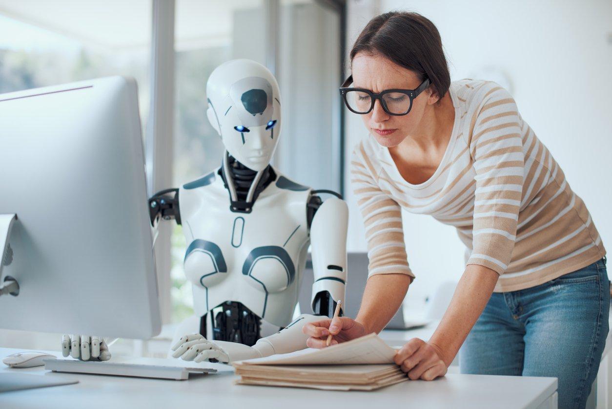 Robô de Inteligência Artificial sentado em uma cadeira de frente uma mesa de escritório com uma mulher ao lado. Ambos estão olhando para umas folhas apoiadas na mesa e fazendo anotações.