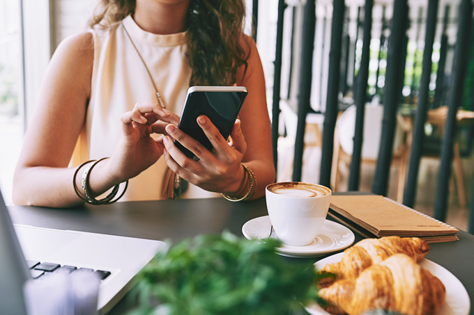 Mulher usando smartphone sentada em uma mesa com um notebook, um café e pães