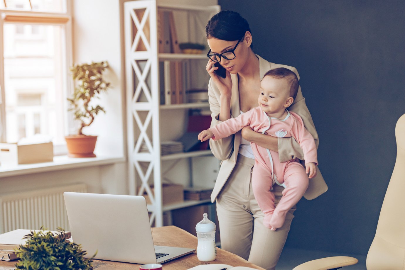 Mãe empreendedora segura bebê enquanto fala ao telefone e usa o computador: trabalho de casa, home office