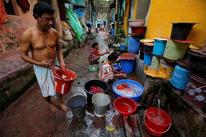 Índia: homem coleta água potável enquanto mulheres lavam suas roupas e utensílios, em uma torneira municipal na favela de Calcutá.