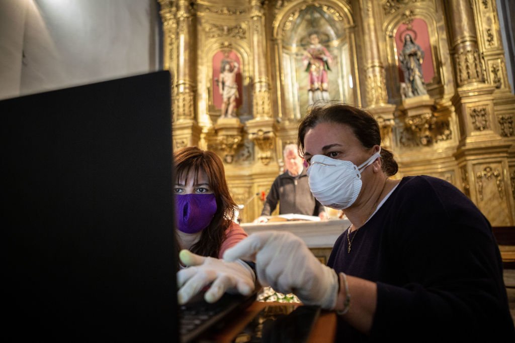 Duas mulheres usando máscara ajustam computador para transmissão de missa pela internet, na Espanha