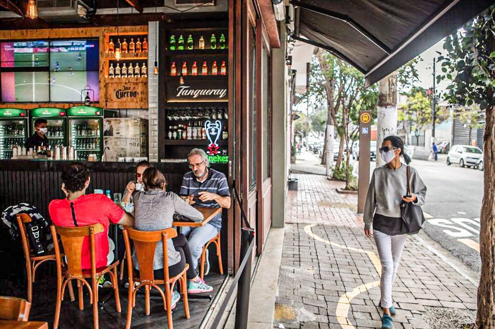 Pedestre usando máscara protetora passa por pessoas sentadas em um restaurante no bairro Vila Madalena, em São Paulo. Segunda-feira, 6 de julho de 2020.