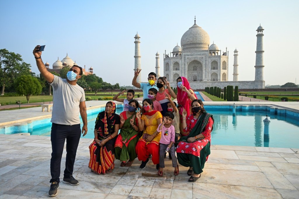 Um grupo de pessoas, de máscara, posam para selfie em frente ao Taj Mahal, castelo na Índia com piscina em frente